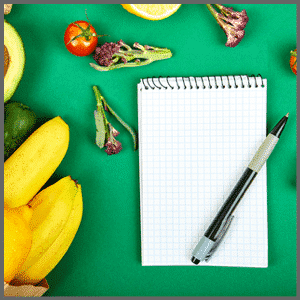 Leeg notitieblok met een pen naast bananen, avocado, tomaat en sla op een groene ondergrond.