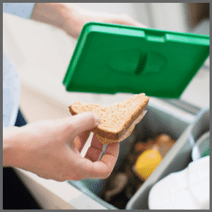 Een hand houdt een snee brood boven een open afvalbak met etensresten en een groen deksel.