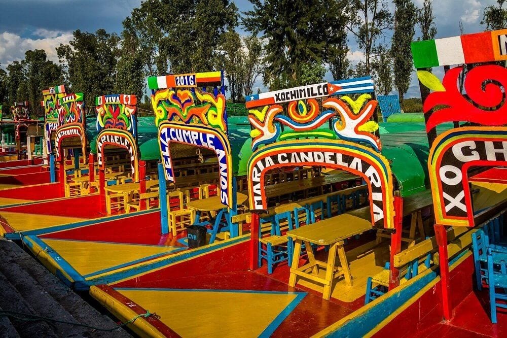 Trajineras de colores en Xochimilco, con arcos decorados y mesas rodeadas de sillas de madera.