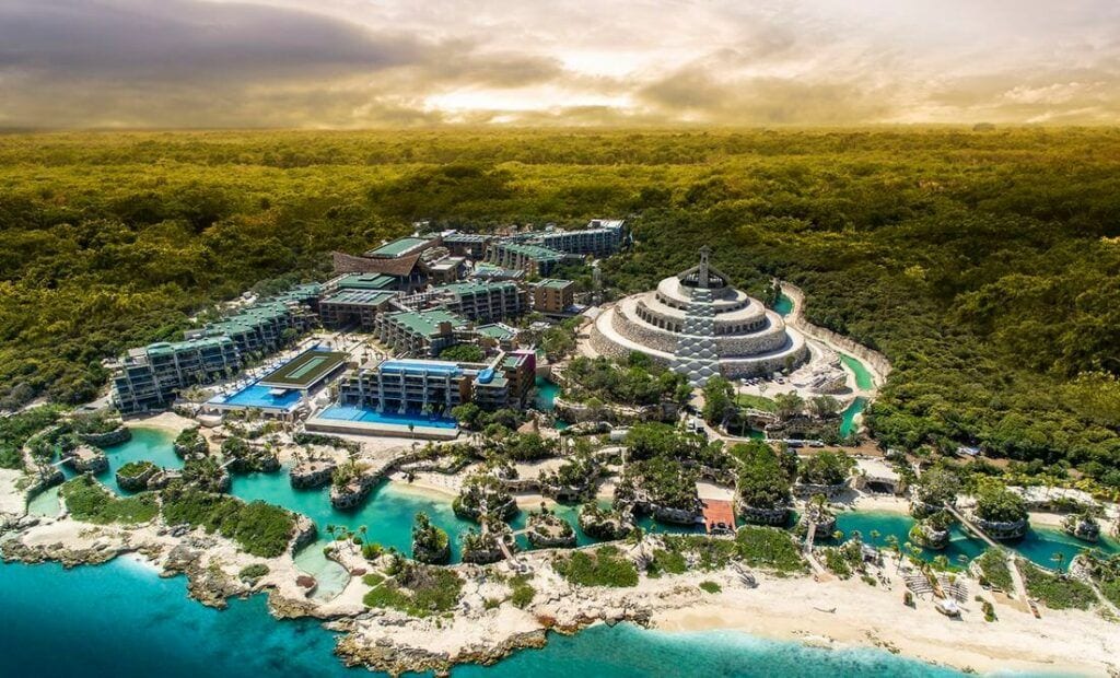 Vista aérea de Xcaret en Playa del Carmen, con edificios rodeados de selva, canales de agua turquesa y playa.