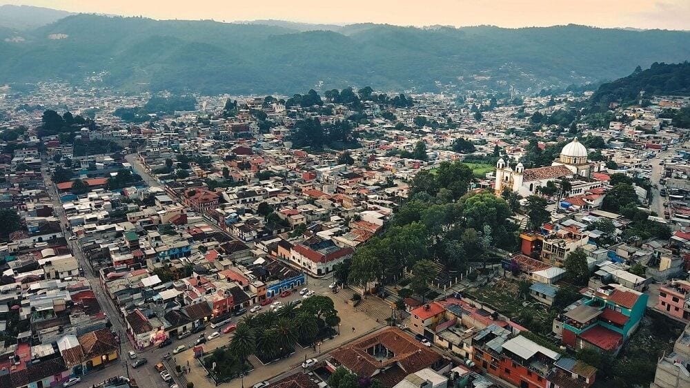 Vista aérea de San Cristóbal de las Casas, Chiapas, con una plaza arbolada, una iglesia y montañas al fondo.