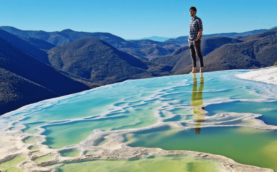 Hombre de pie en una poza de agua turquesa con formaciones minerales en Hierve el Agua, Oaxaca, rodeada de montañas.