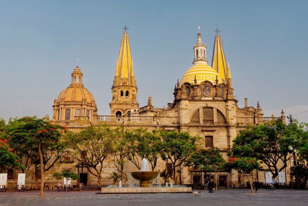 Catedral de Guadalajara, con sus torres amarillas y cúpulas, frente a una plaza con árboles y una fuente.