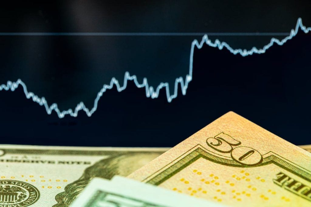 Yhdysvaltain dollarin seteleitä ja taustalla näytöllä vaihteleva, kokonaisuutena nouseva kurssikäyrä.