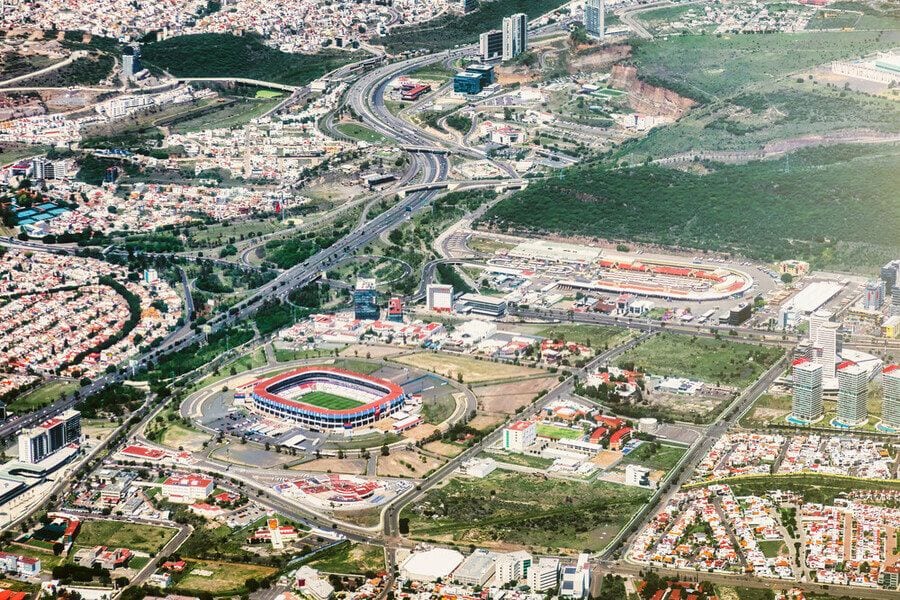 Vista aérea de Querétaro con una autopista, zonas residenciales, edificios modernos y un estadio de futbol rodeado de cerros