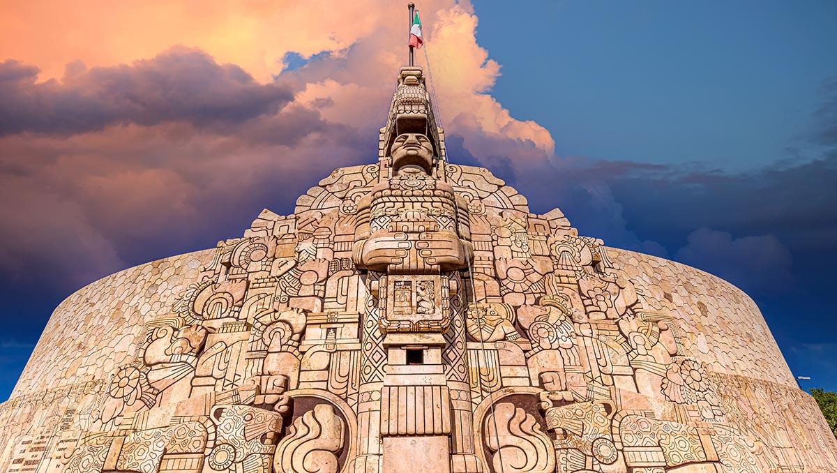 Monumento a la Patria en Mérida, Yucatán, con relieves mayas de piedra y la bandera de México bajo un cielo con nubes al atardecer