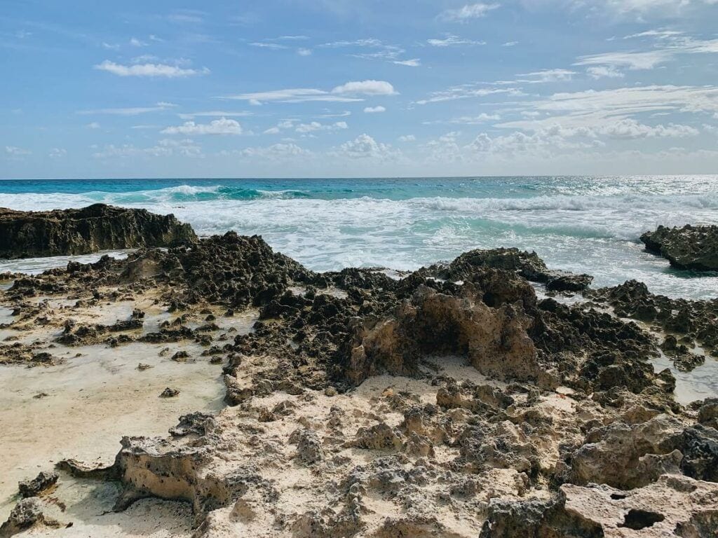 Costa rocosa de Cancún con pequeñas pozas, oleaje blanco y mar turquesa bajo un cielo con nubes dispersas.